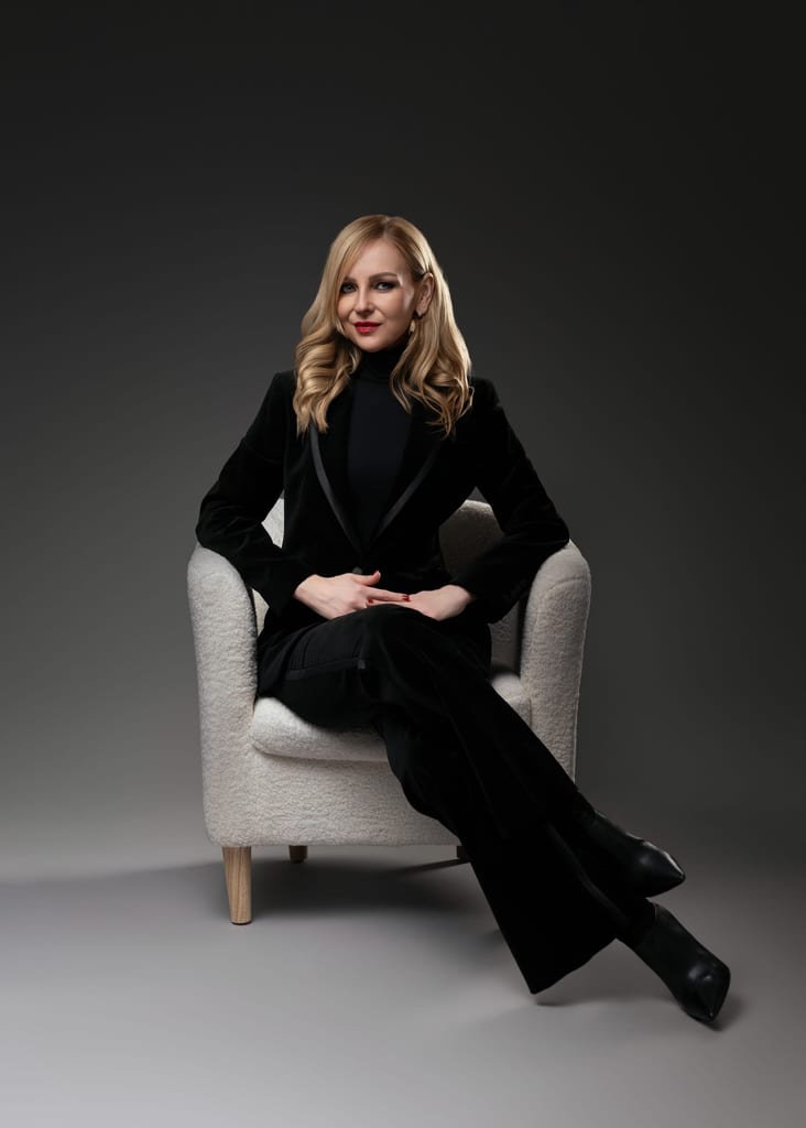 Dr Kamila Szuberska in a black velvet suit, seated in a white armchair