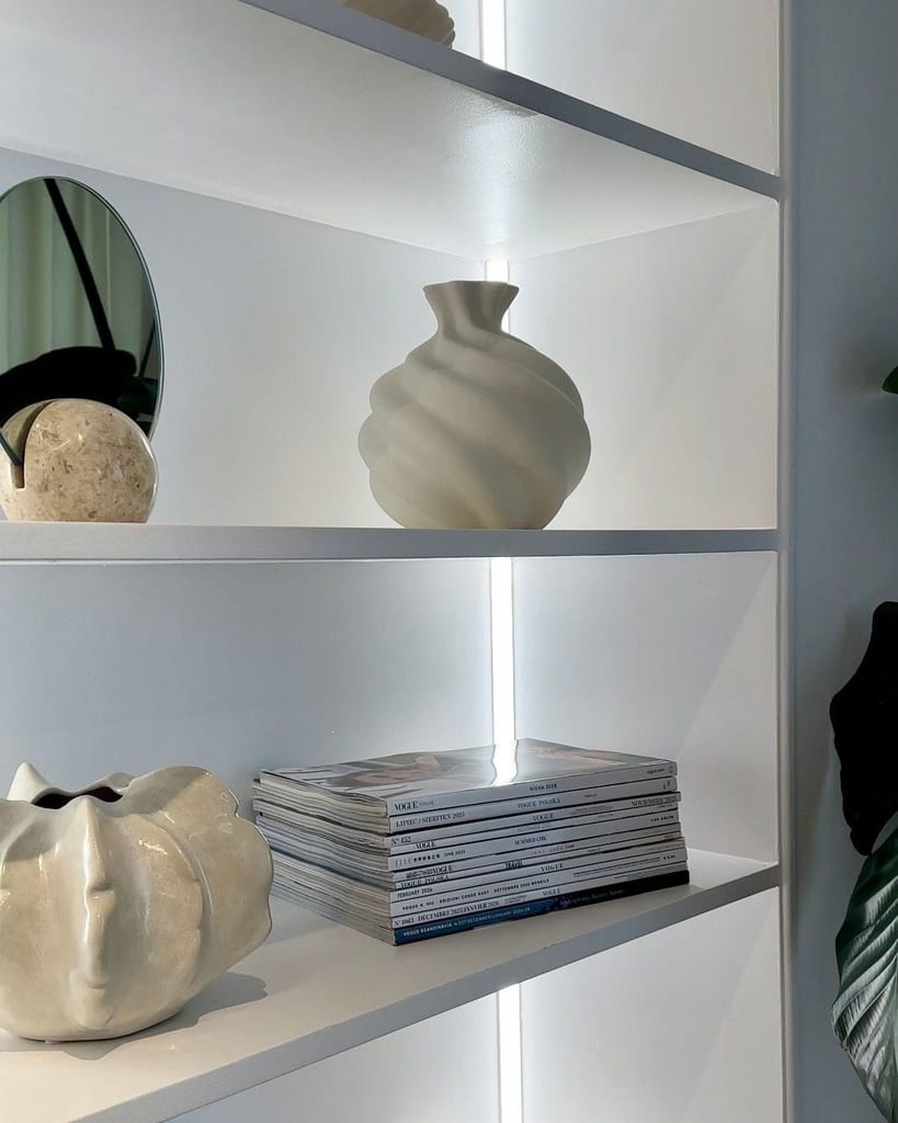 Illuminated white shelves with sculpted cream vases, a round mirror and stacked magazines.