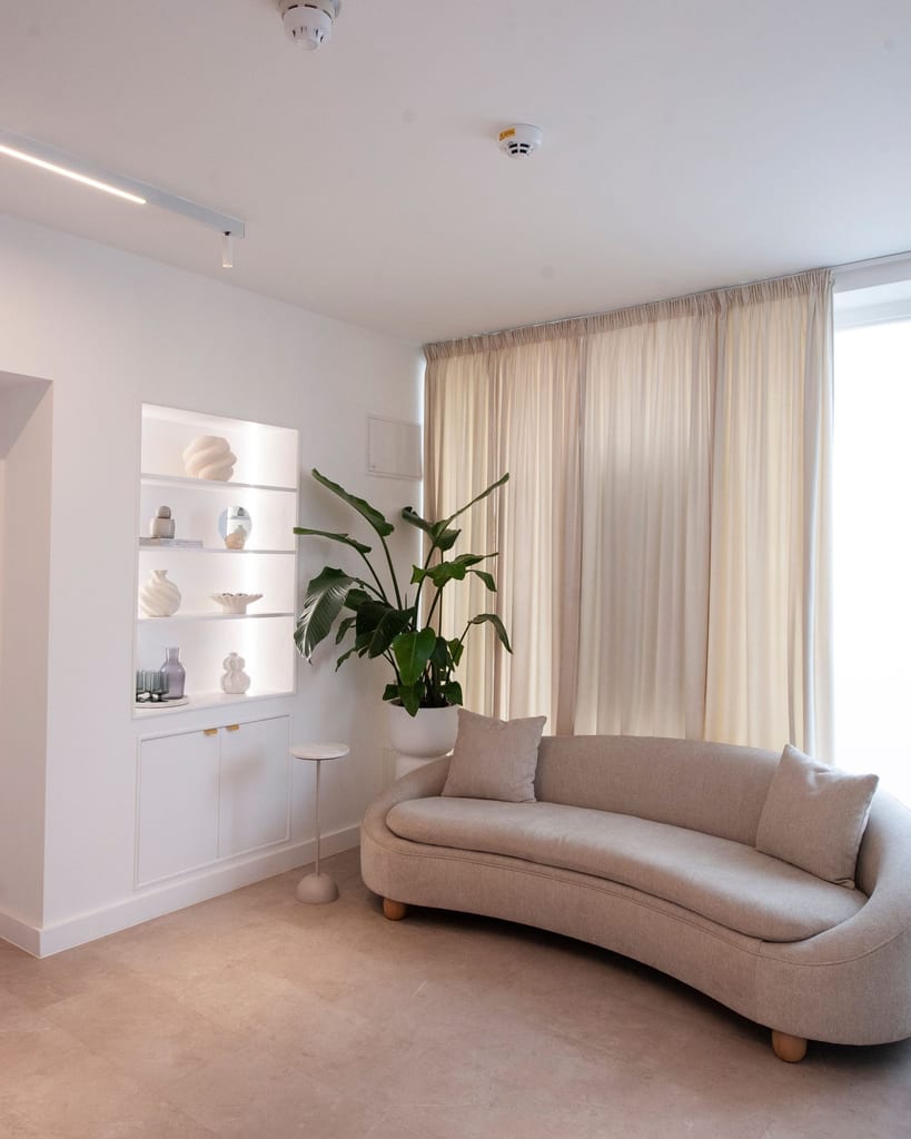 Waiting area with a curved oatmeal sofa, tall plant, illuminated ceramics and floor-length curtains.